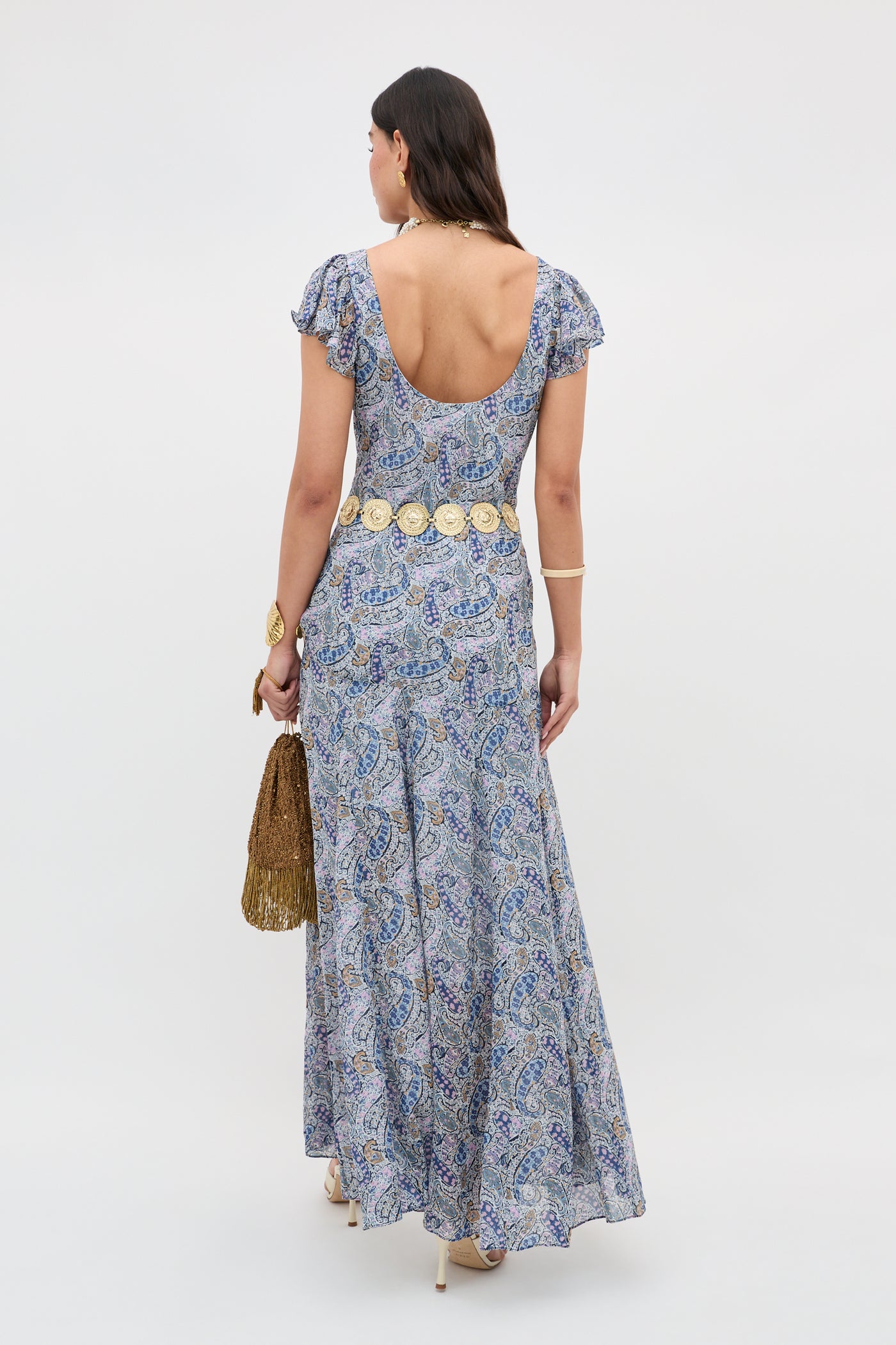 A woman faces away, wearing the Erianne - Bourton Navy dress in blue and white Liberty fabric with a paisley print. She accessorizes with a gold fringed bag, decorative belt, and bracelets.