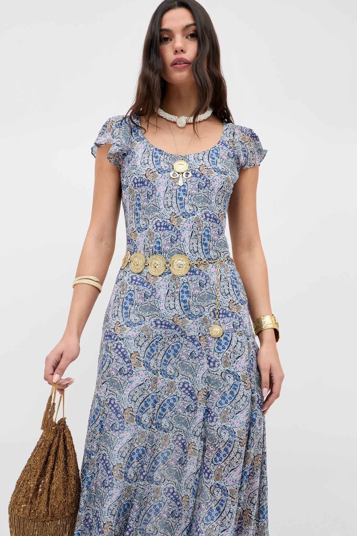 A woman wears the Erianne - Bourton Navy paisley print georgette dress with gold jewelry and a brown woven bag, standing against a plain light background.