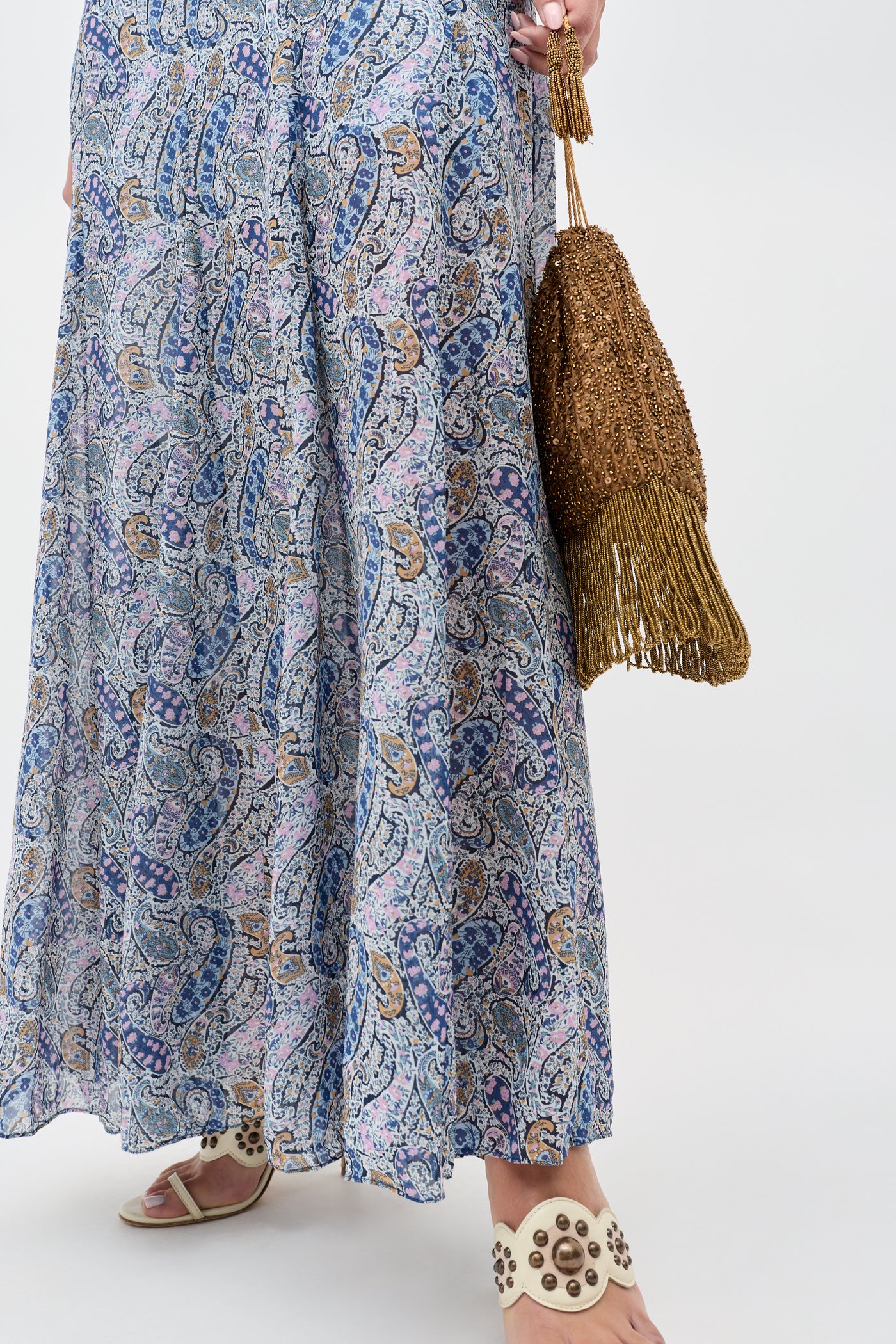 Wearing the Erianne - Bourton Navy long blue and white paisley print skirt, white studded sandals, and holding a brown fringed woven bag against a plain white background.