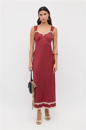A woman with dark hair stands against a white background wearing the Lucie - Rose sleeveless, floor-length red dress with light lace trim. She carries a black textured handbag with a scarf, gold sandals, and jewelry.