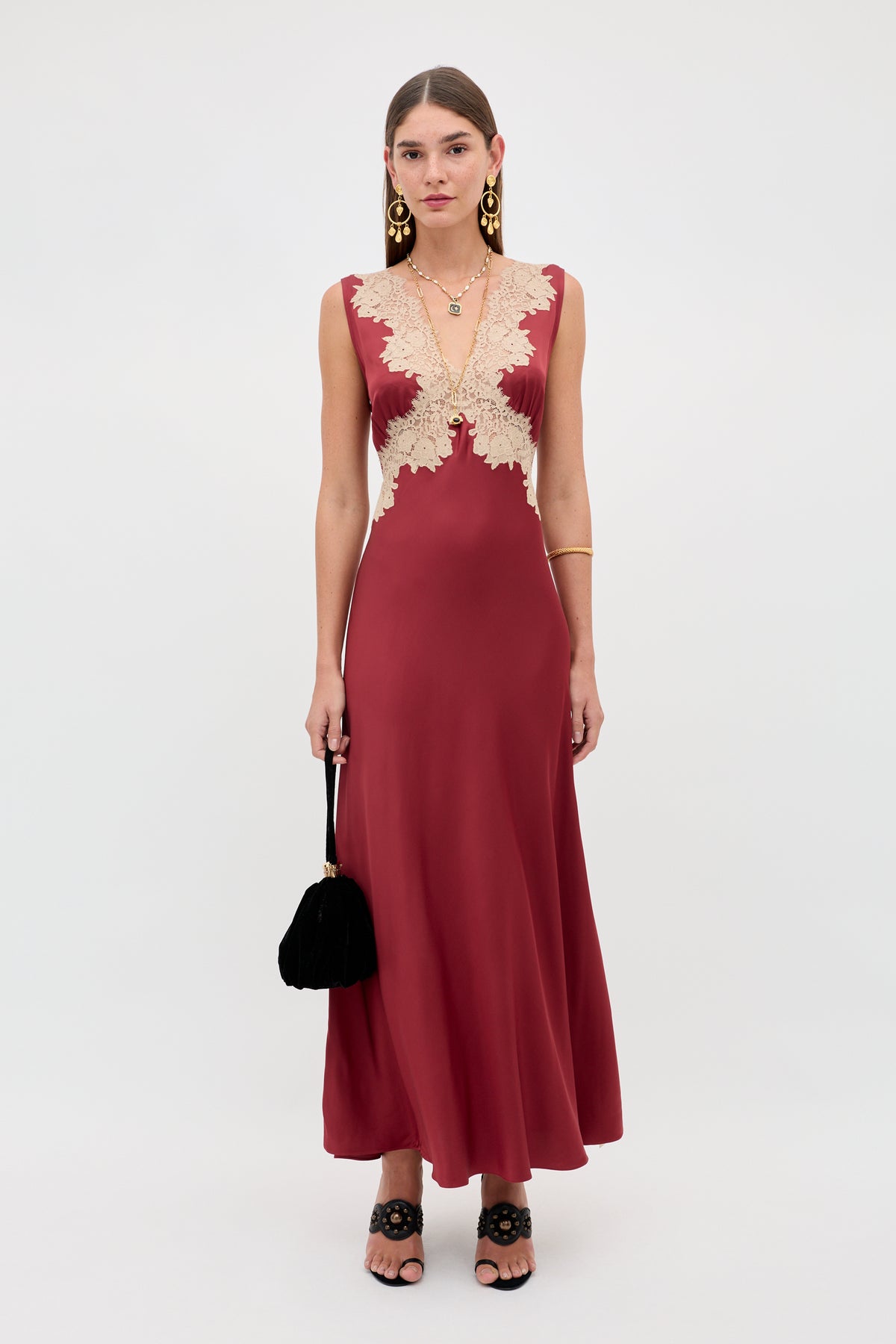 A woman stands against a plain background wearing the Lysandra - Rose dress, a sleeveless long burgundy gown with beige lace bodice detailing. She accessorizes with gold earrings, a small black handbag, and has straight hair.