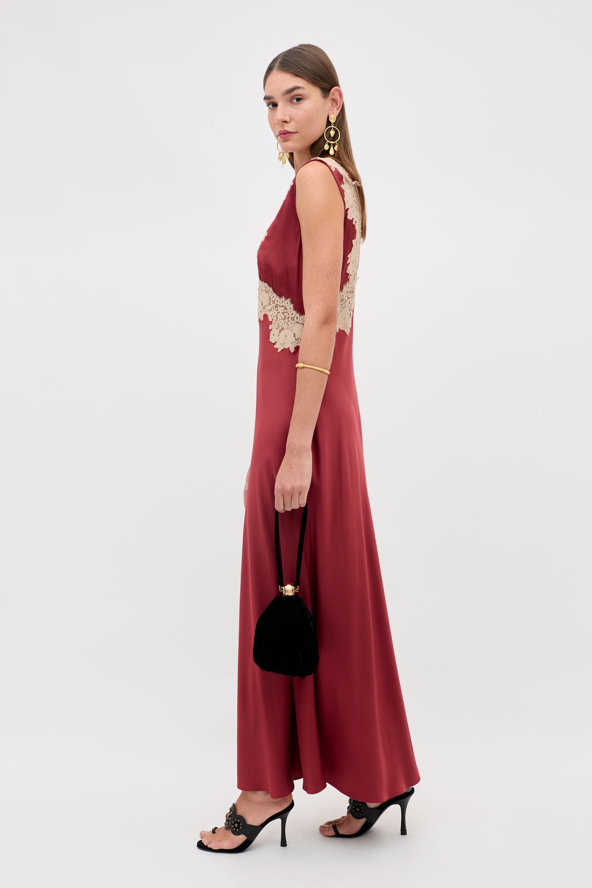 A woman in the Lysandra - Rose sleeveless, ankle-length maroon dress with beige lace accents stands sideways, holding a black handbag. She pairs it with high-heeled sandals, gold earrings, a bracelet, and wears her straight hair down.