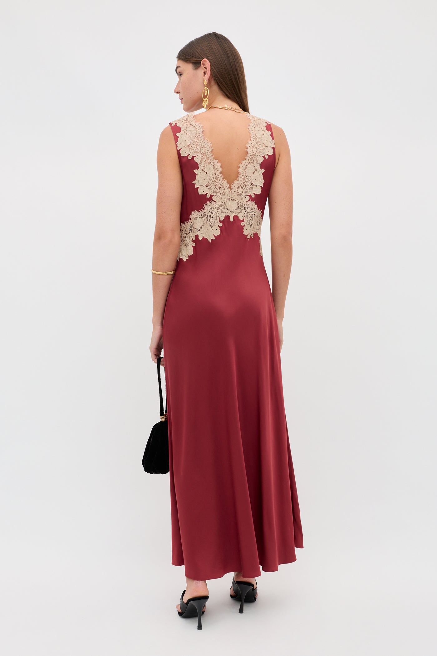 A woman in the Lysandra - Rose sleeveless, long burgundy satin dress with beige lace back stands facing away. She carries a small black handbag, wears black heels, gold hoop earrings, and a bracelet.
