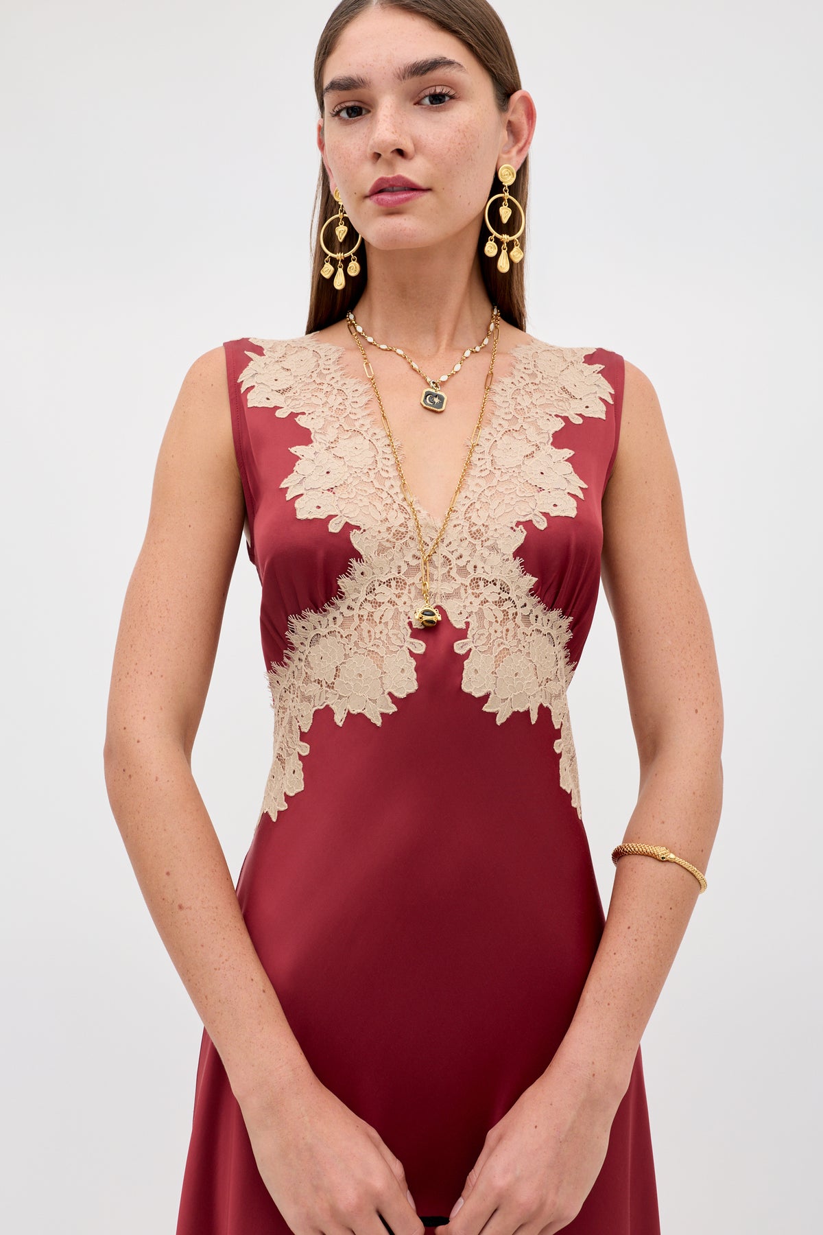 A woman stands against a plain light background wearing the Lysandra - Rose sleeveless burgundy dress with beige lace bodice details, gold earrings, layered necklaces, and a bracelet.