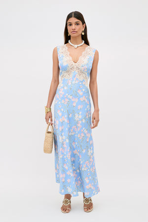 A woman wearing the Lysandra - Watercolour Floral Blue dress, paired with a straw handbag, statement jewelry, and heeled sandals, stands against a plain light background.