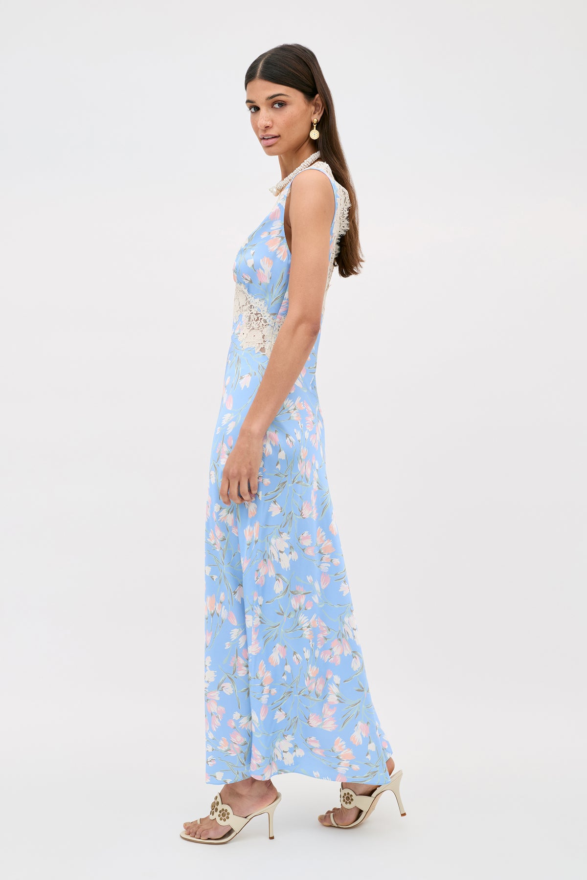 A woman stands in profile against a plain light background, wearing the Lysandra - Watercolour Floral Blue sleeveless maxi dress with pink florals, lace waist detail, gold earrings, and cream high-heeled sandals.