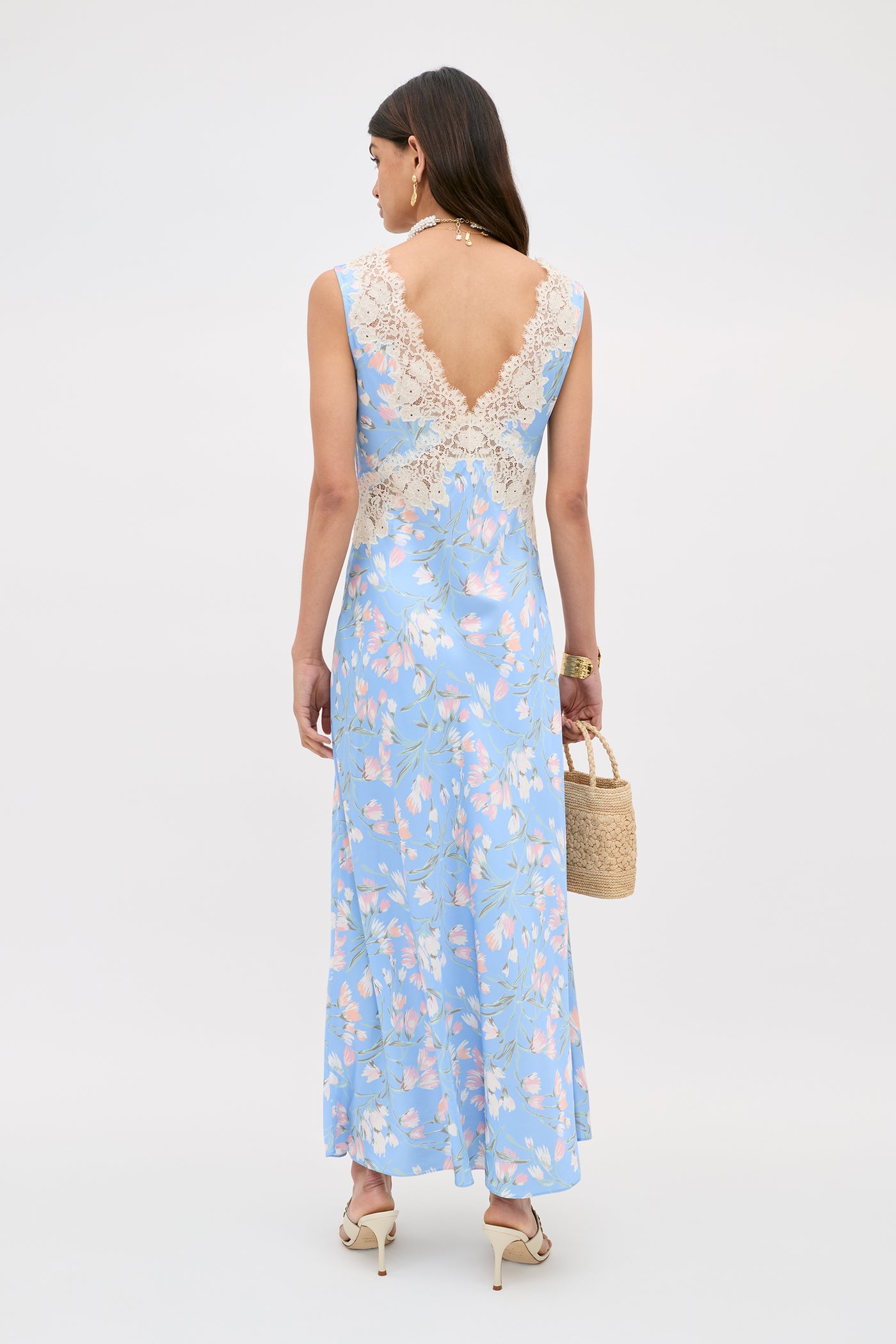 A woman in the Lysandra - Watercolour Floral Blue sleeveless dress featuring a deep V-back with lace trim stands facing away, holding a woven straw handbag and wearing heels.