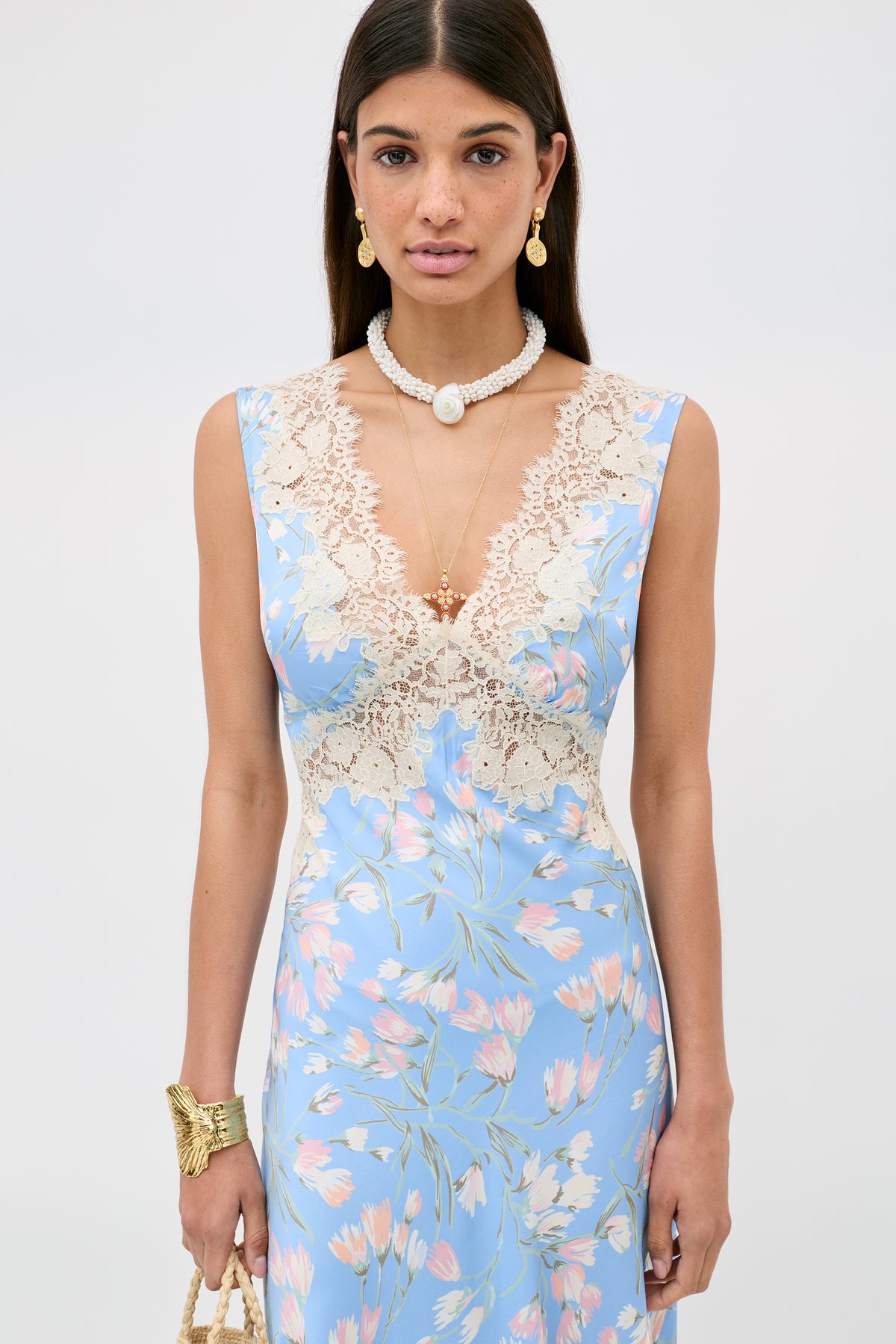 A woman in the Lysandra - Watercolour Floral Blue sleeveless dress with a deep V lace neckline, styled with layered necklaces, pearl earrings, a gold bracelet, and a woven handbag poses against a plain background.