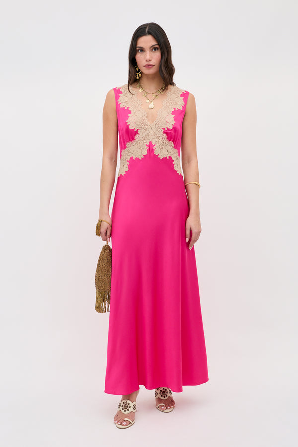 A woman stands against a plain white background wearing the Lysandra - Cerise sleeveless, floor-length hot pink dress with beige lace bodice detailing, holding a gold tasseled clutch and accessorized with gold jewelry.