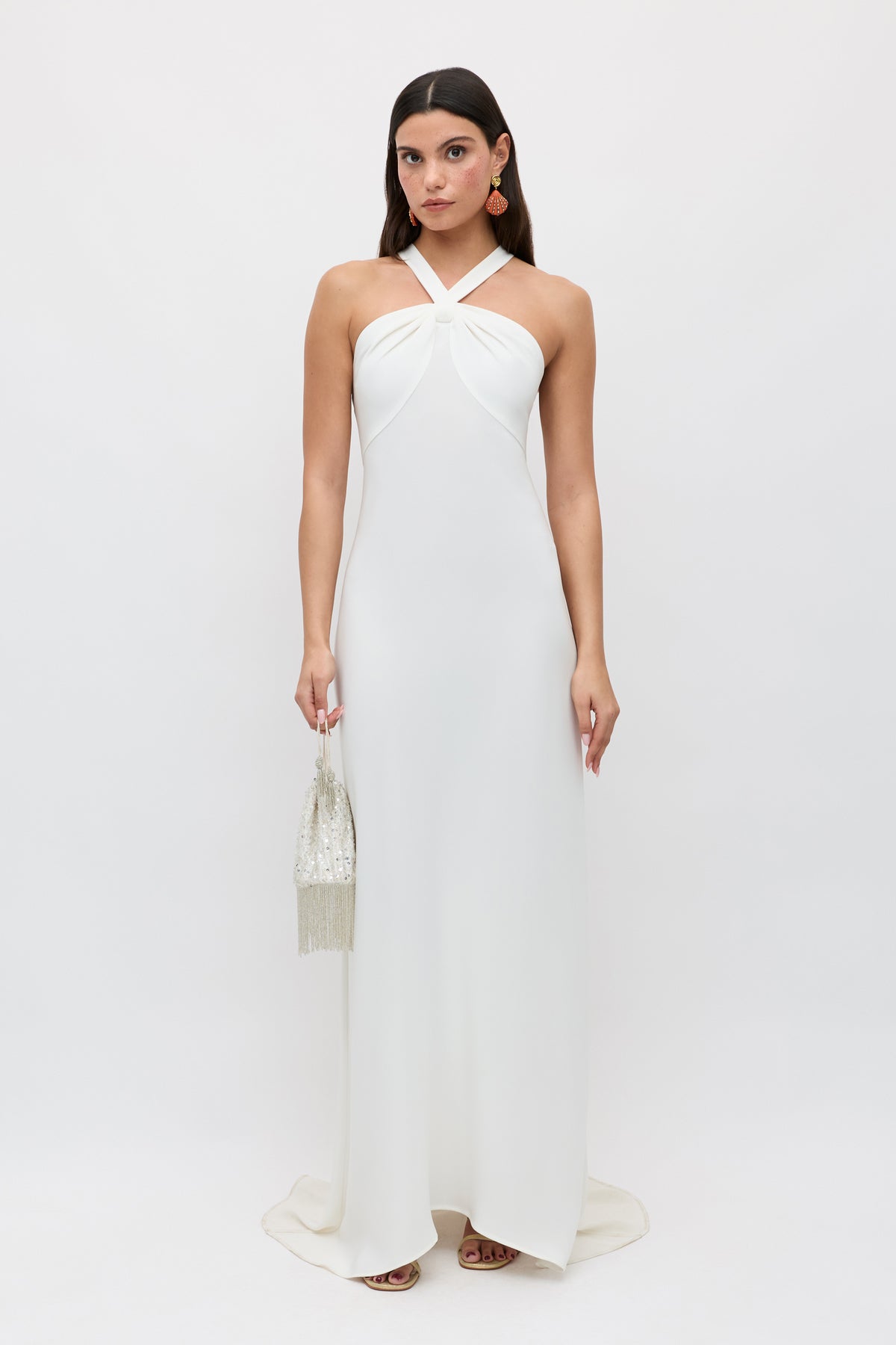 A woman stands against a white background wearing the Giorgia - Ivory, a long twist neck halter bridal gown in satin back crepe. She holds a small beaded handbag, wears dangling earrings, and her dark hair is styled straight and loose.