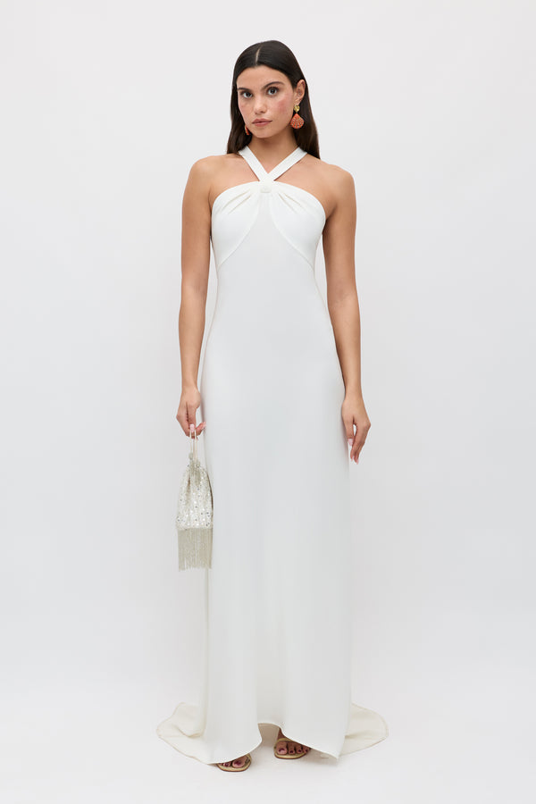 A woman stands against a white background wearing the Giorgia - Ivory, a long twist neck halter bridal gown in satin back crepe. She holds a small beaded handbag, wears dangling earrings, and her dark hair is styled straight and loose.