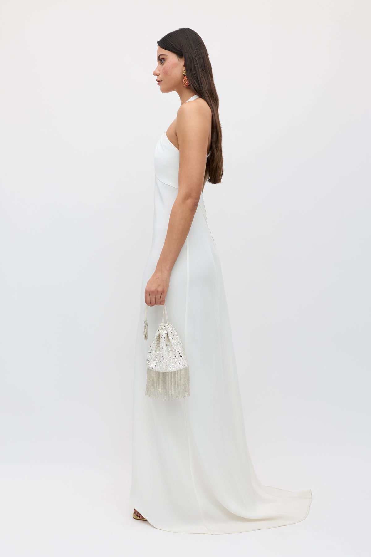 A woman in the Giorgia - Ivory twist neck halter bridal gown stands in profile against a plain background, holding a beaded white fringe purse. Her long dark hair is down and she wears drop earrings.