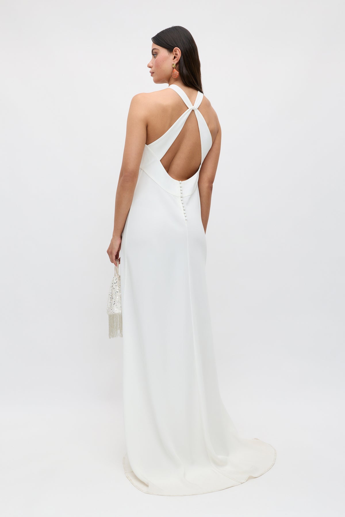 A woman in the Giorgia - Ivory long satin back crepe bridal gown stands facing away, showcasing its open back with crisscross straps. She holds a small beaded bag, her straight dark hair down, all set against a plain white background.