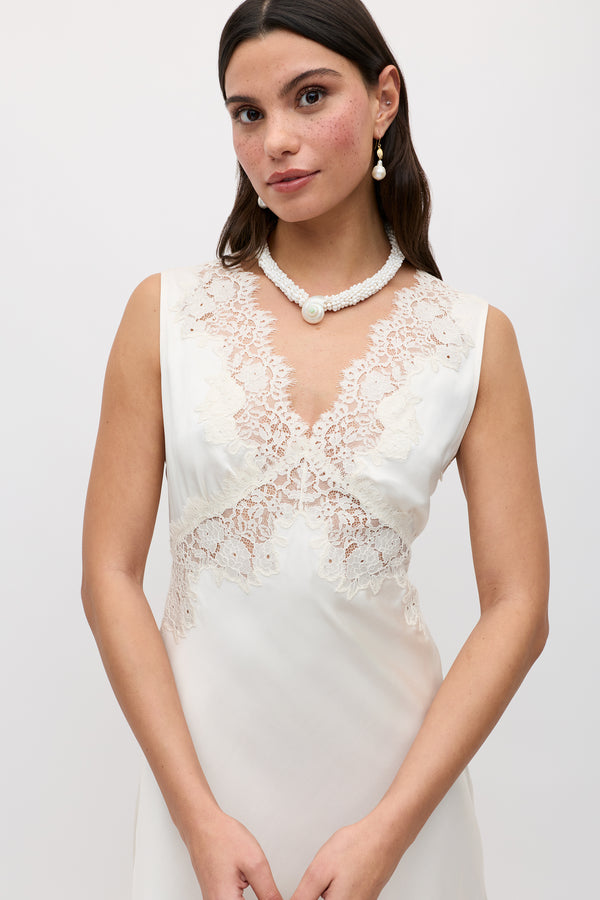 A woman in the Azzurra - Pearl sleeveless vintage slip dress with lace at the chest and waist, accessorized with pearl earrings and a white beaded necklace featuring a large pearl, stands against a plain light background.