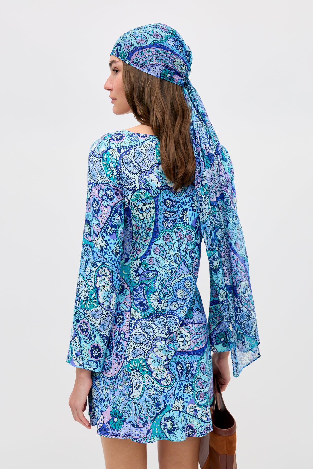 A woman in the Julien - Paisley Floral Blue dress and matching headscarf stands facing away, holding a brown bag against a plain light background.