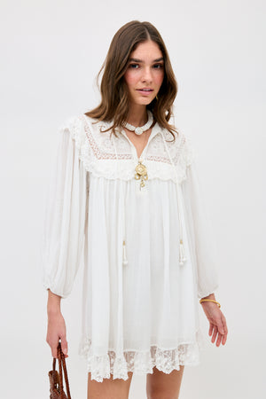 A woman in the Inez - Lilly Embroidery Ivory dress, featuring loose long sleeves and lace trim, accessorized with layered necklaces and a brown woven bag, stands against a plain background.