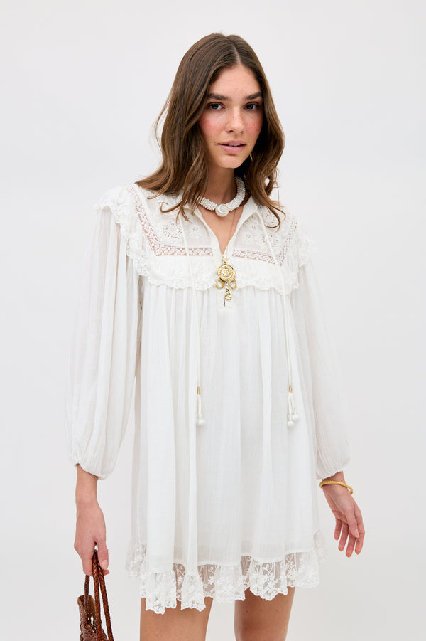A woman in the Inez - Lilly Embroidery Ivory dress, featuring loose long sleeves and lace trim, accessorized with layered necklaces and a brown woven bag, stands against a plain background.