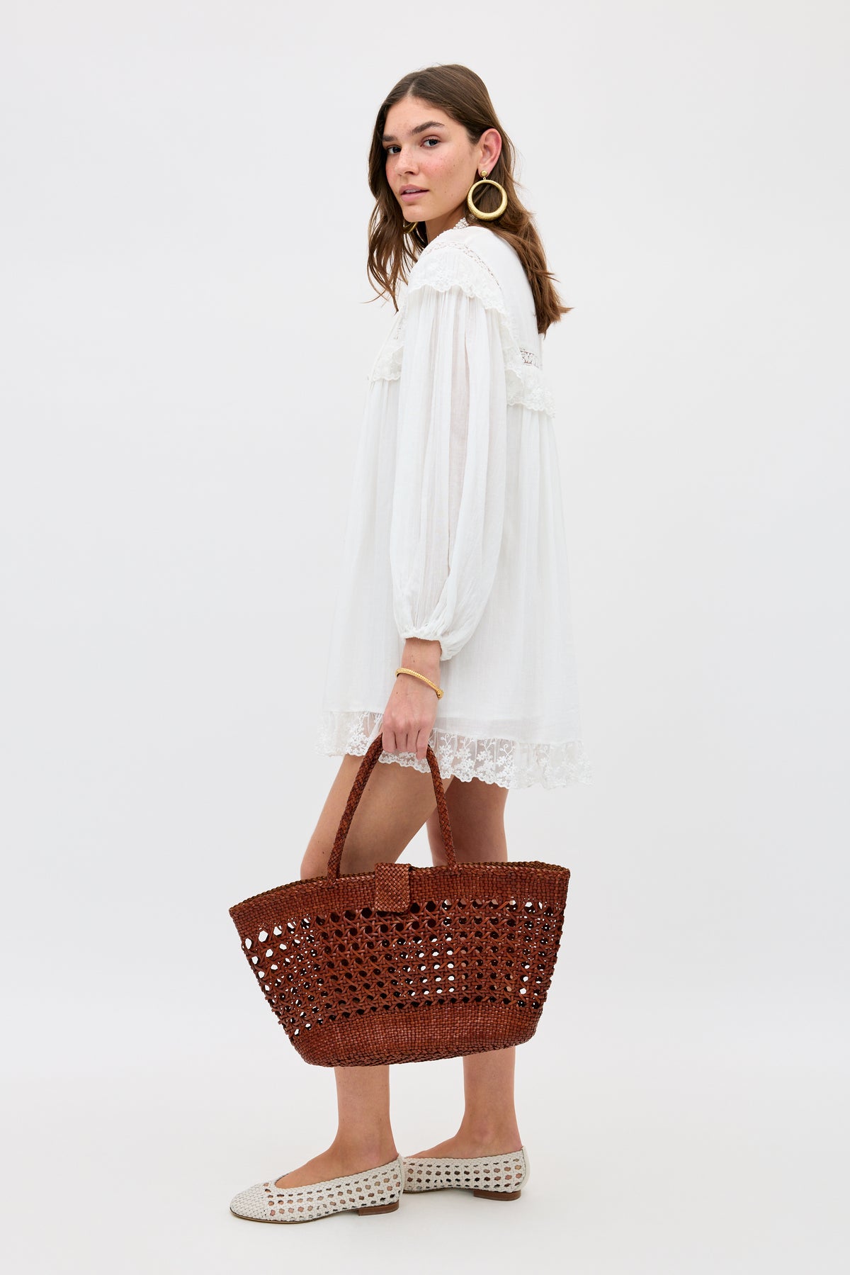 Wearing the Inez - Lilly Embroidery Ivory, a woman with wavy brown hair stands sideways, accessorized with large hoop earrings. She pairs the loose embroidered dress with white woven flats and holds a large brown woven tote.