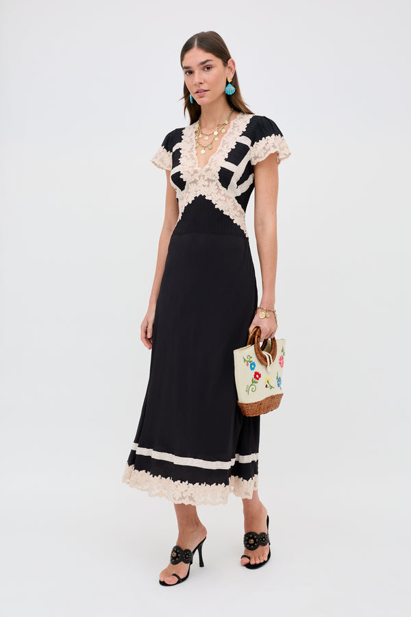 A woman stands wearing the Martina - Black midi dress with beige lace details, black heeled sandals, turquoise earrings, gold layered necklaces, and holding a beige woven handbag featuring colorful floral embroidery.