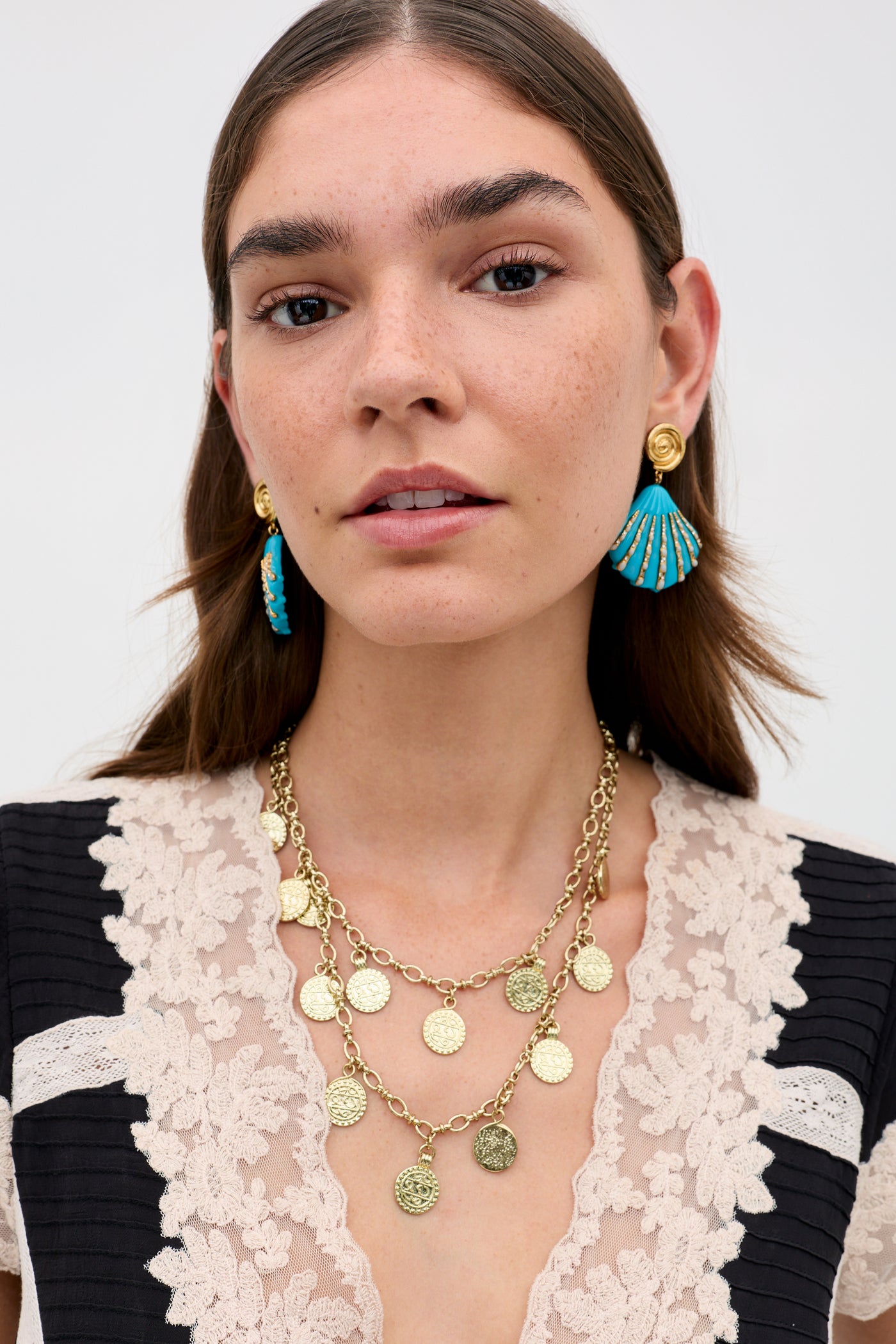A woman with long brown hair wears gold coin jewelry—a layered necklace and blue-and-gold earrings. She models the Martina - Black top with cream lace detailing, looking directly at the camera.