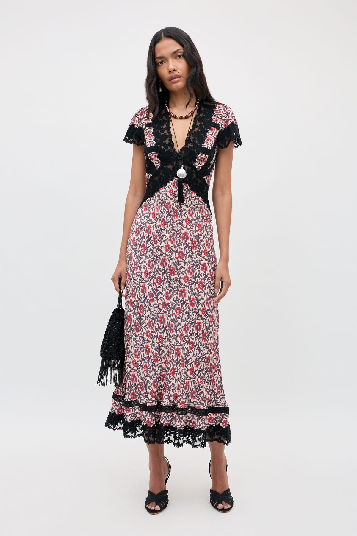 A woman wearing the Martina - Sarasa Rose Red floral midi dress with black lace trim, styled with black heels and a fringed black bag, stands against a plain light background.