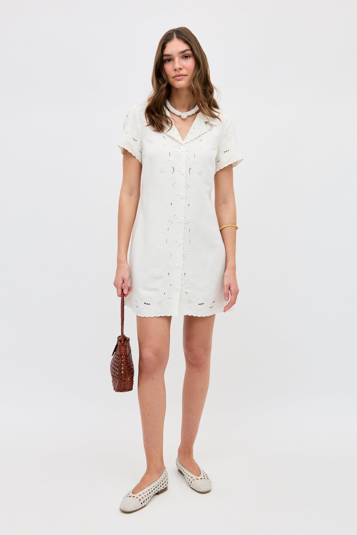 A woman stands against a plain white background wearing the Zayanna - Esther Cutwork Embroidery Ivory dress, white woven shoes, a beaded necklace, a gold bracelet, and holding a brown textured handbag.