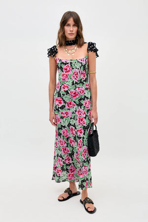 A woman stands against a white background wearing Adele - Rose Dot Mix Black, a long floral dress with pink flowers and green leaves, paired with black sandals, a black polka dot choker, and holding a black textured handbag.