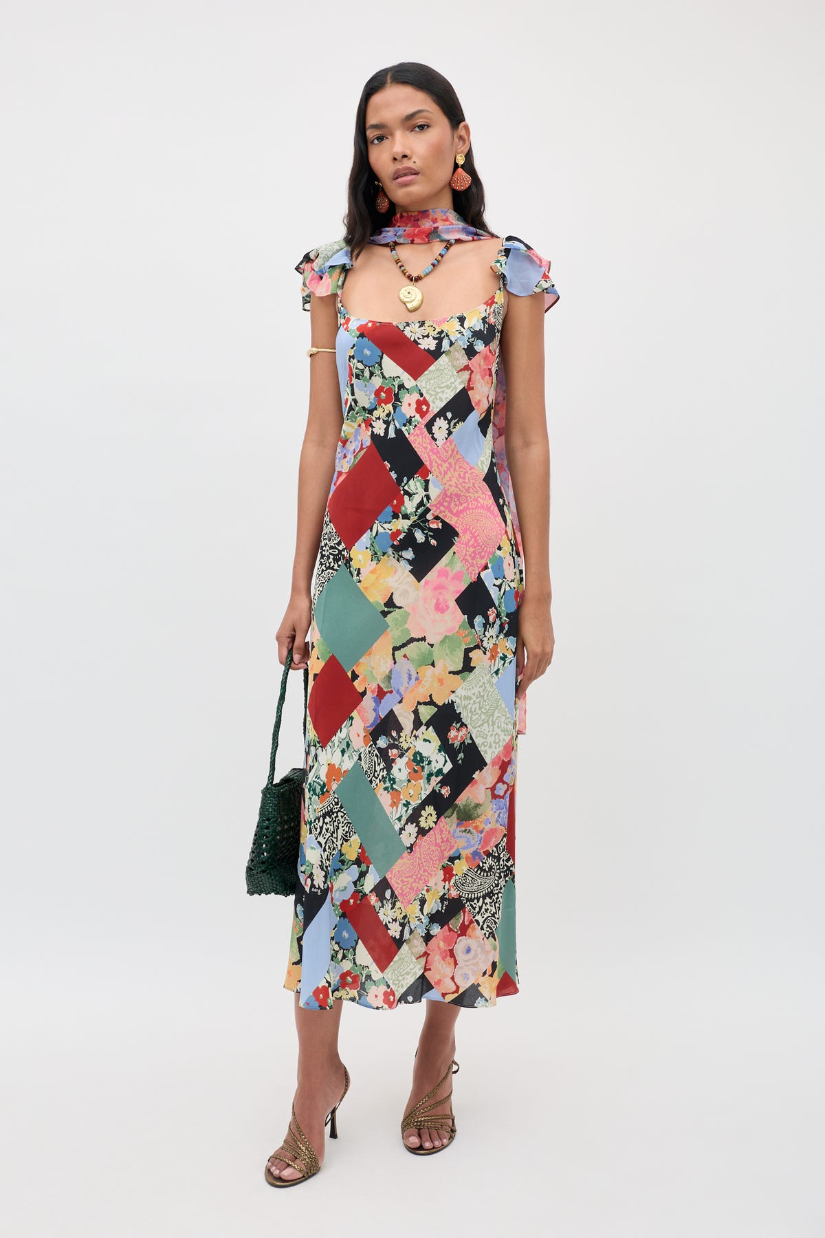 A woman wearing the Adele - Olivia Patchwork Multi midi dress with ruffled sleeves, paired with a matching scarf and sandals, holding a dark green handbag and standing against a plain white background.