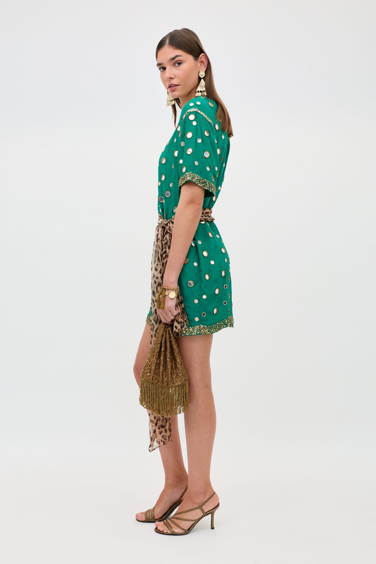Here’s a rewritten sentence using the provided product data: A woman in the Cruz - Lume Embellishment Dark Green dress, featuring a patterned short silhouette with gold accents, accessorized with gold earrings and bracelets, holding a fringed brown bag and wearing heeled sandals, stands against a plain white background.