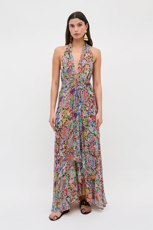 Woman wearing the Alaina - Cara Floral Leopard Brown, a sleeveless halter-neck floor-length dress with a colorful abstract floral and leopard pattern. She stands against a white background, hair down, in black sandals and gold statement earrings.