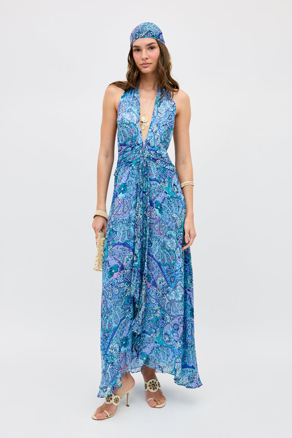 A woman poses against a plain background in the Alaina - Paisley Floral Blue maxi dress, featuring a flowing silhouette and deep V-neck. She accessorizes with a matching headscarf, bangles, necklace, heeled sandals, and a small woven bag.