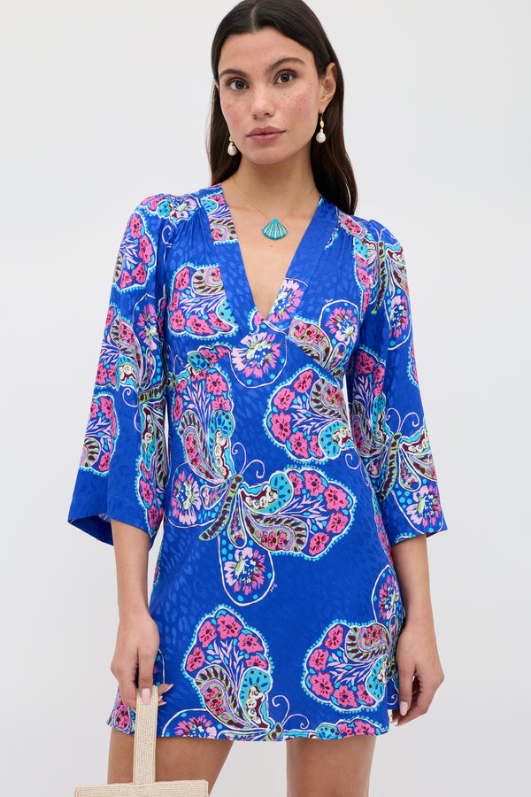A woman wearing the Margaux - Butterfly Bloom Jacquard Blue dress, featuring colorful paisley patterns, three-quarter sleeves, and a V-neckline, holds a beige bag against a plain light background.