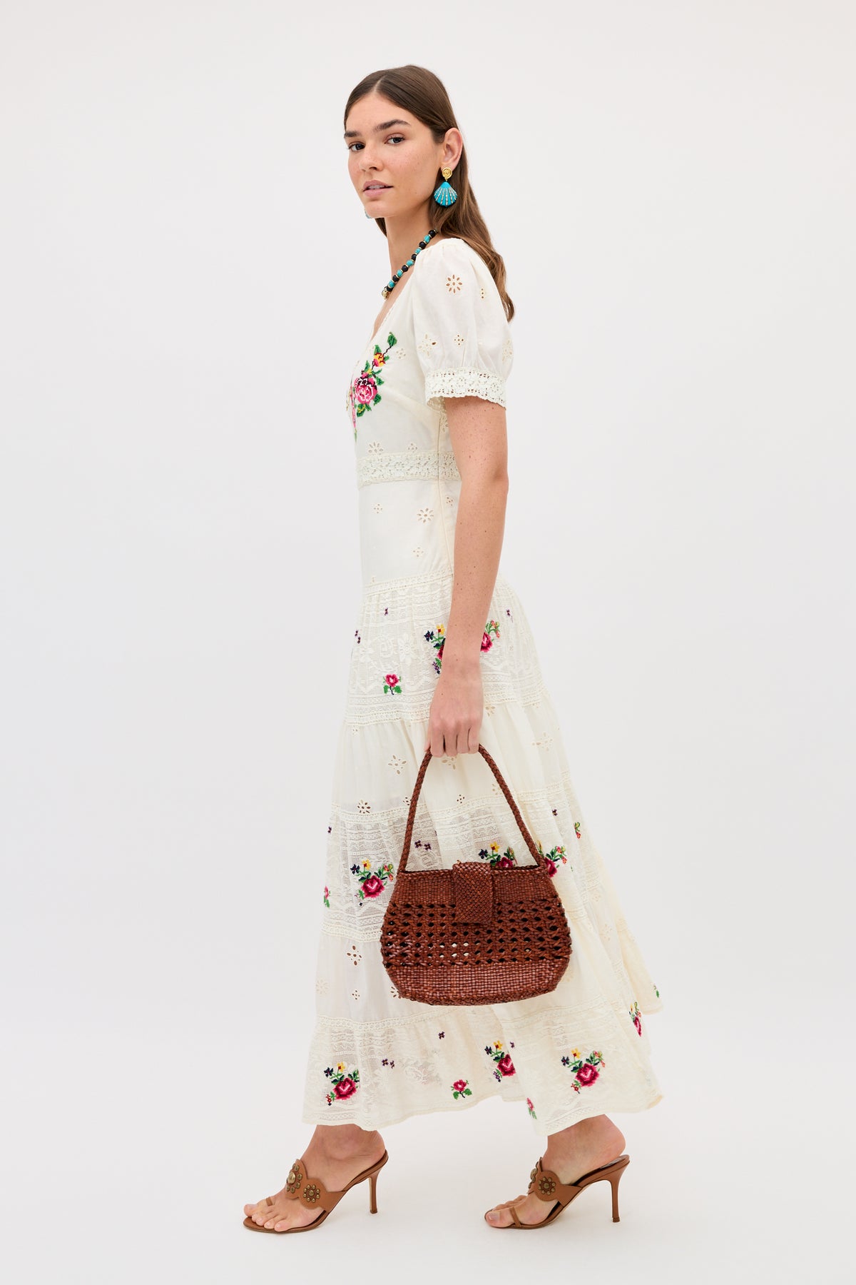 A woman wears the Cady - Spring Rose Embroidery Cream dress, featuring white fabric with colorful floral embroidery. She pairs it with a woven brown handbag, brown heeled sandals, and turquoise earrings against a white background.