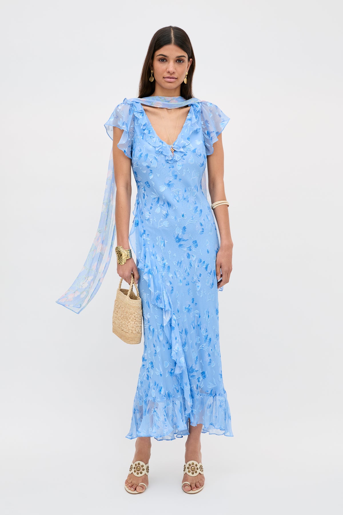 A woman wears the Meadow - Ava Devore Blue floral ruffled midi dress with a matching sheer scarf, styled with a straw handbag, gold jewelry, and beige sandals against a white background.