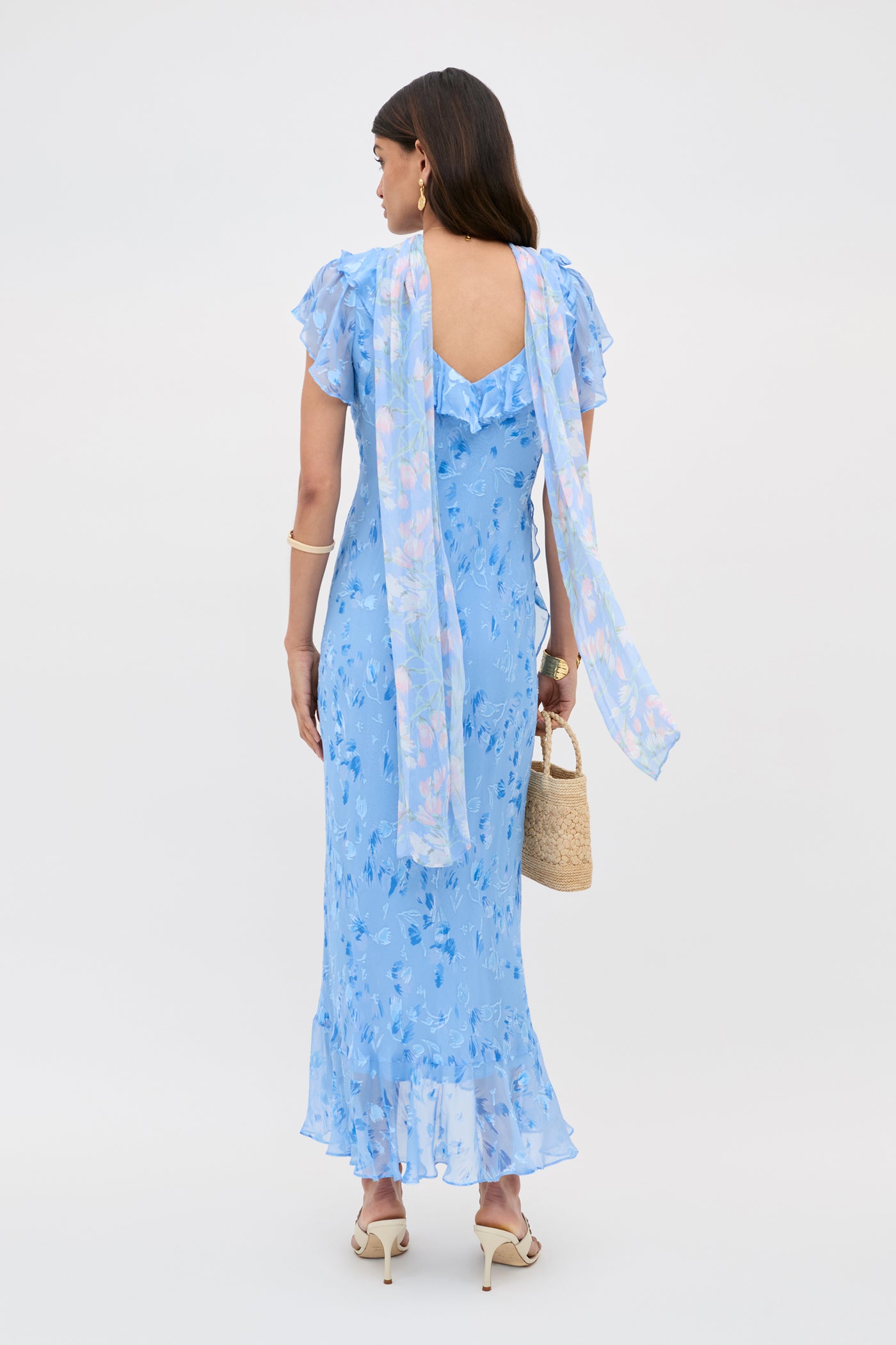 A woman faces away in the Meadow Ava Devore Blue dress, featuring light blue florals, ruffled sleeves and hem, a back keyhole cutout, and a long sheer scarf. She carries a woven bag with white heeled sandals and gold hoop earrings.