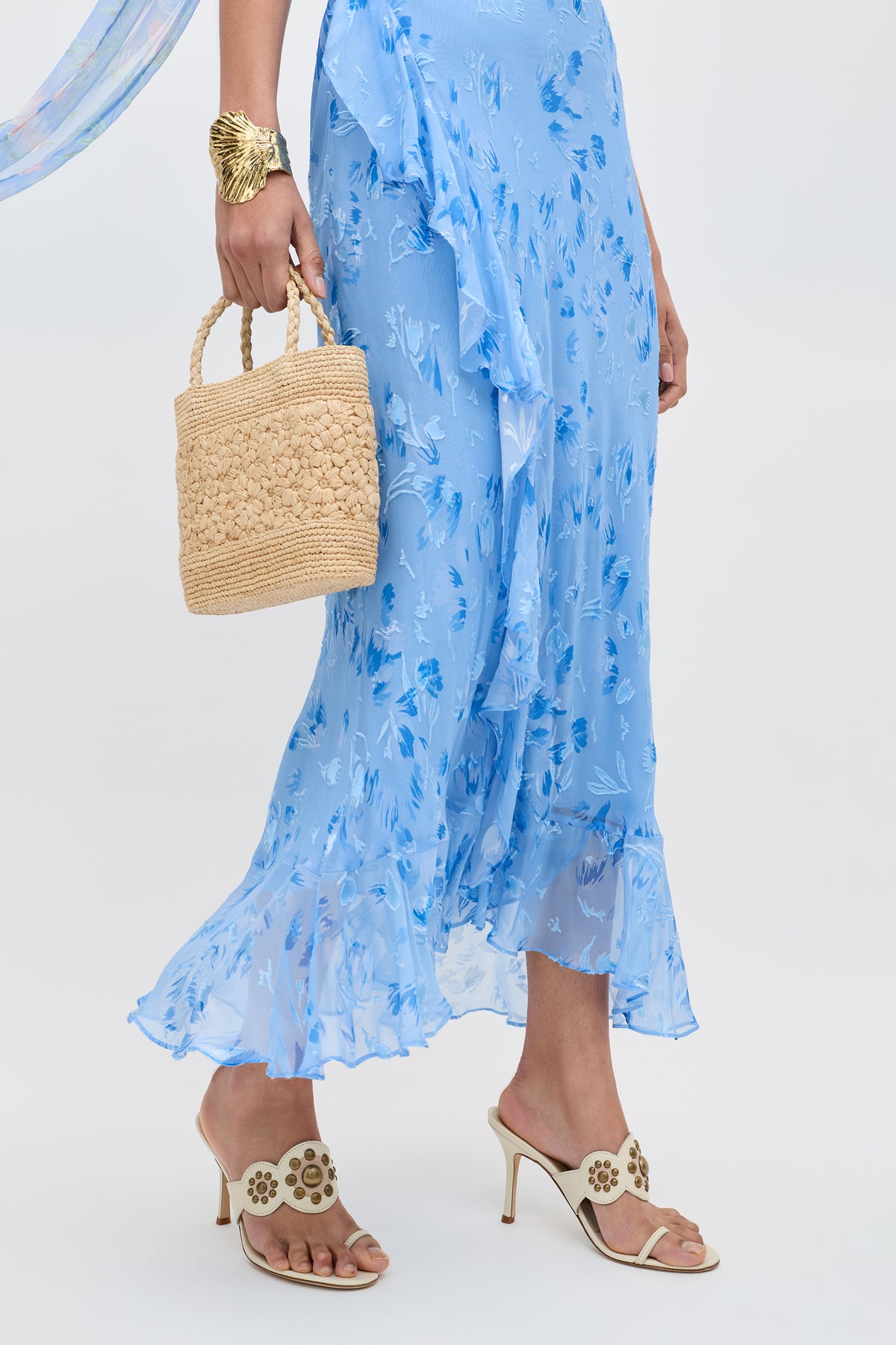 A person wearing the Meadow - Ava Devore Blue dress, beige heeled sandals with floral cutouts, a chunky gold bracelet, and holding a woven straw handbag against a plain light background.