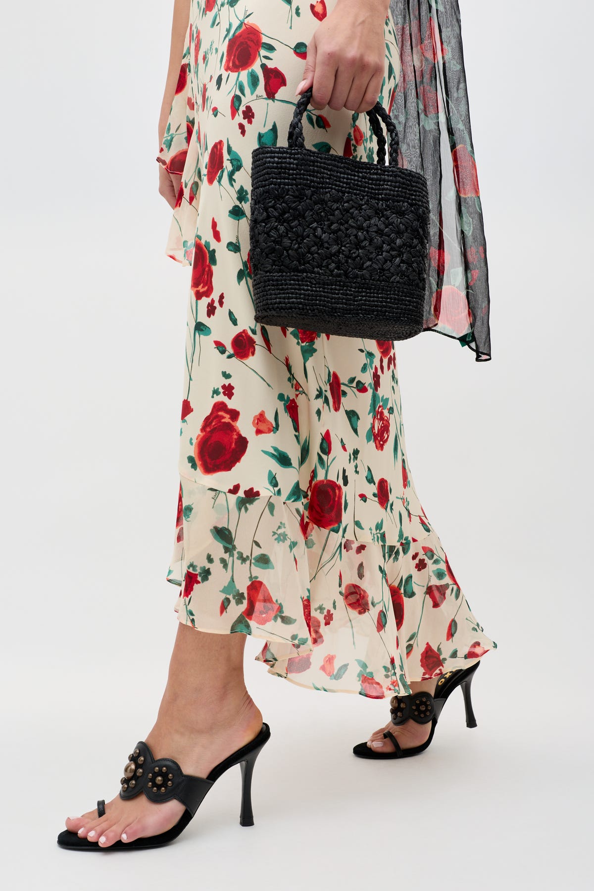 A person wearing the Meadow - Claudia Rose Cream dress with red rose prints and ruffle hem, black high heel sandals, and holding a small black woven handbag; only the lower body and hand are visible.