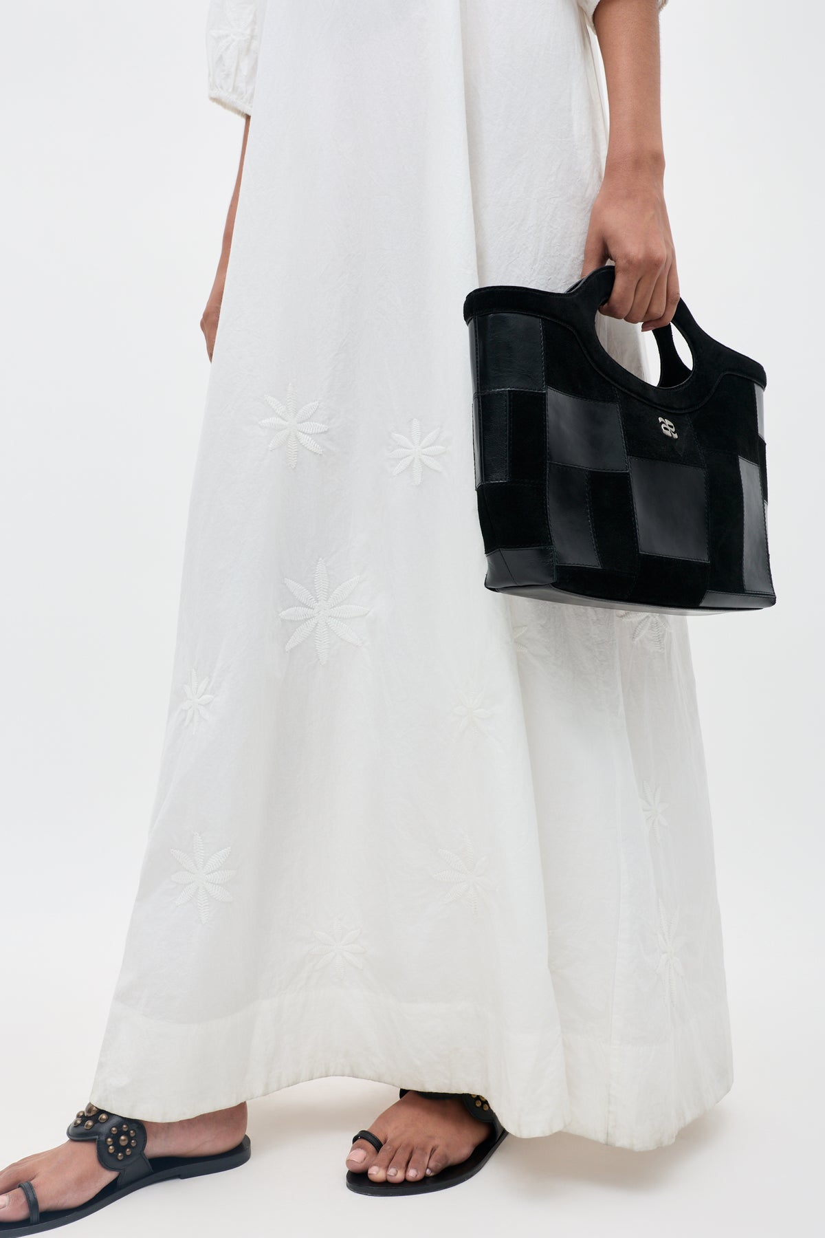 A person is wearing the Gizele - Daisy Floral Embroidery White long dress, holding a black checkered handbag and wearing black sandals. The background is plain and light.