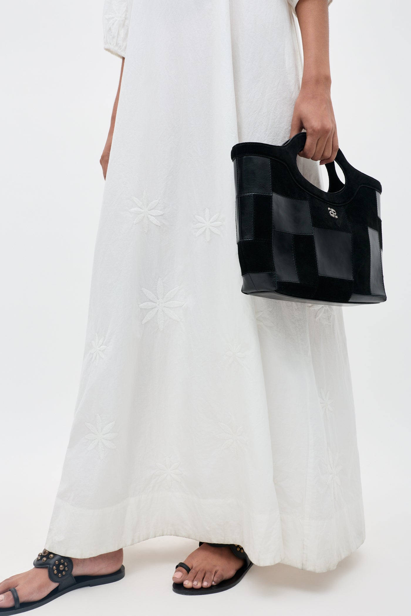 A person is wearing the Gizele - Daisy Floral Embroidery White long dress, holding a black checkered handbag and wearing black sandals. The background is plain and light.