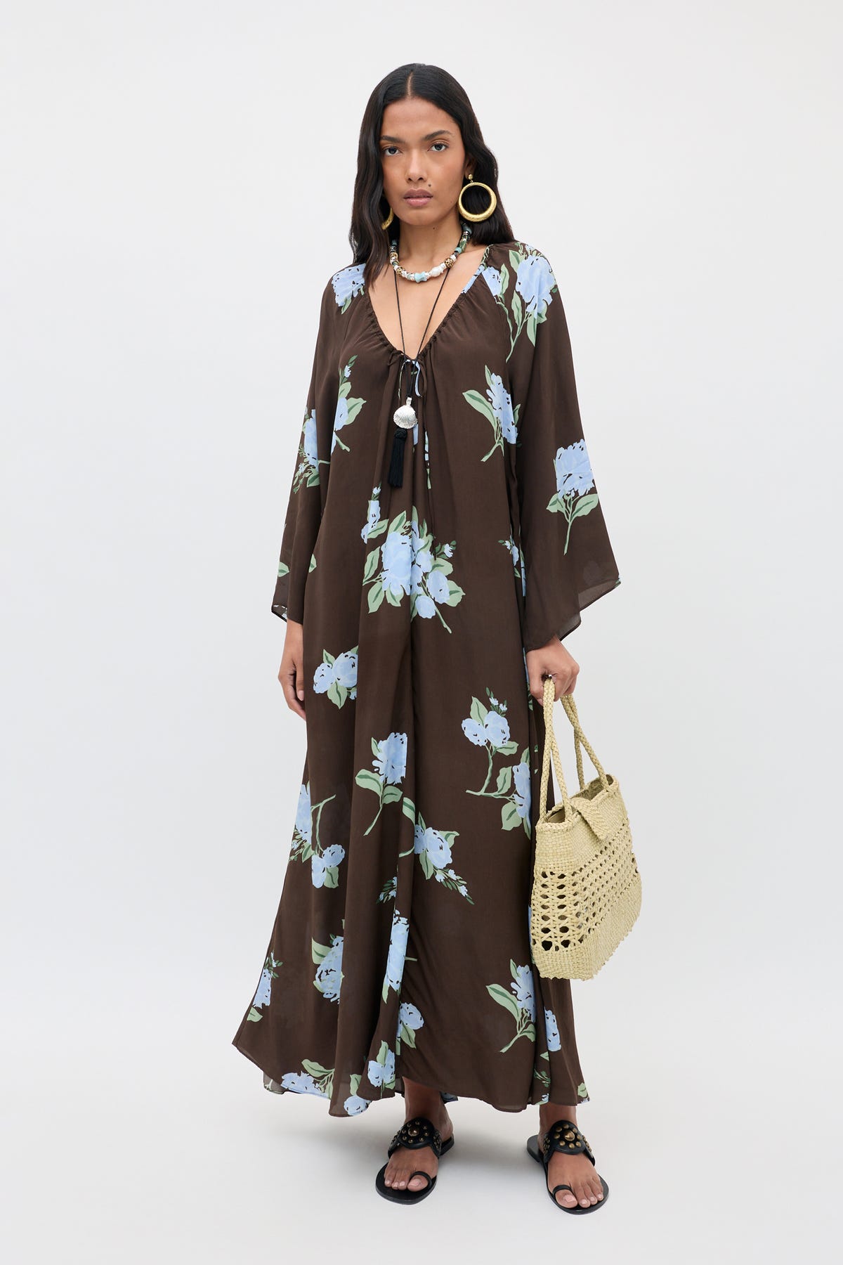 A woman stands against a plain white background wearing the "Lianne - Peonies Scatter Brown" long floral dress with blue flowers, paired with black sandals, layered necklaces, large hoop earrings, and a woven handbag.