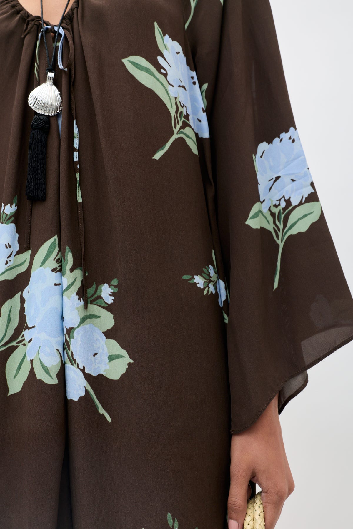 A person is shown holding a woven beige item while wearing the Lianne - Peonies Scatter Brown dress, which features a brown base with blue peony flowers and green leaves, adorned with black and silver tassels. Only their lower face, hand, and upper body are visible.