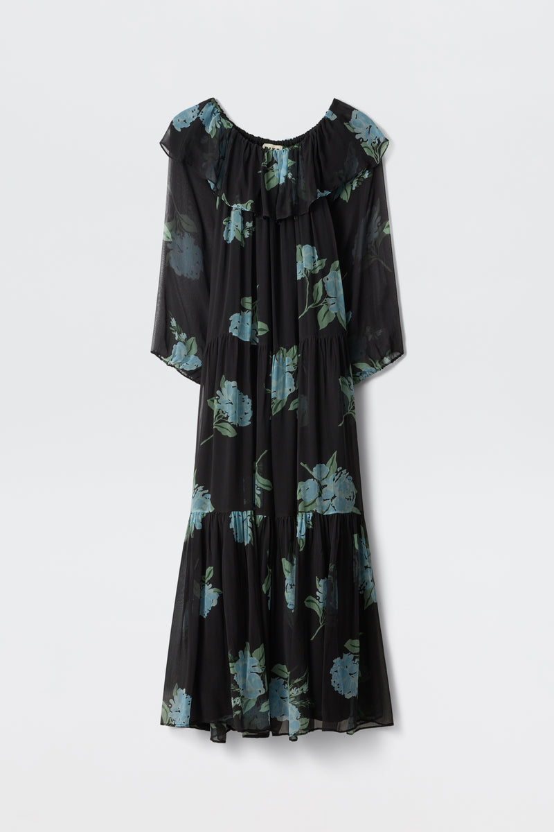 A long-sleeved, ruffled maxi dress in charcoal, featuring blue peony scatter prints and a ruffled off-the-shoulder neckline, is displayed against a plain white background. This dress is named "Lela - Peonies Scatter Charcoal.