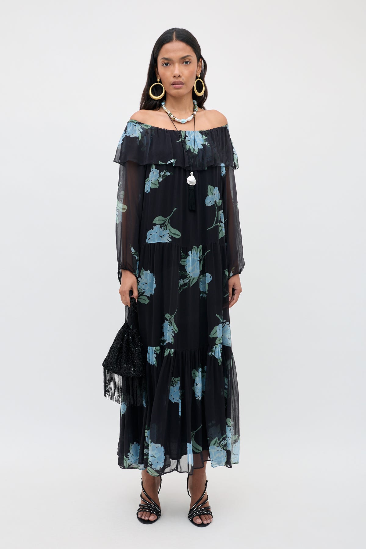 A woman models the Lela - Peonies Scatter Charcoal dress, featuring a black off-the-shoulder silhouette with blue floral print and sheer long sleeves, paired with large hoop earrings, layered necklaces, a black fringe handbag, and strappy black heels against a plain light background.