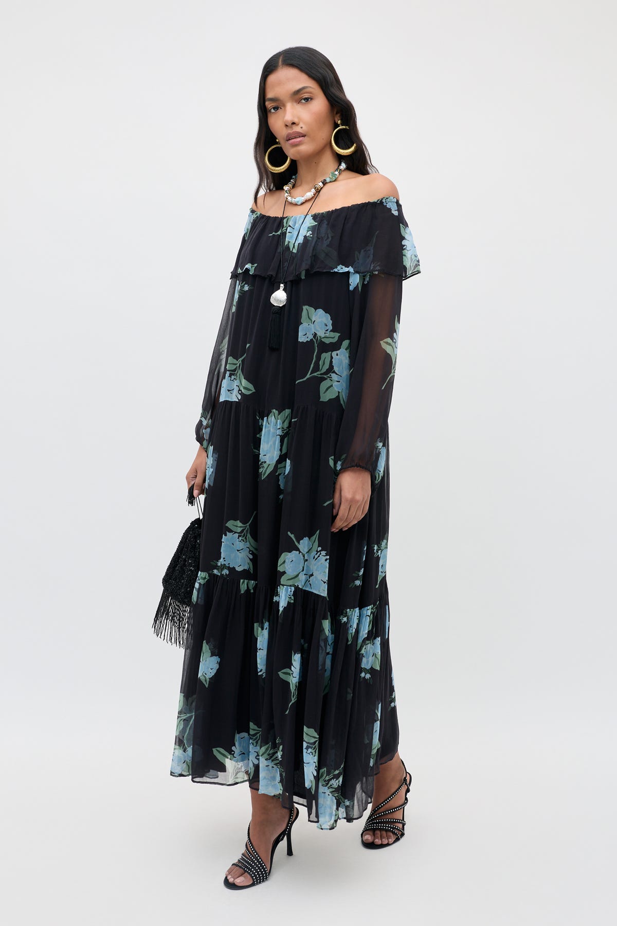 A woman wears the Lela - Peonies Scatter Charcoal, a long, off-the-shoulder black floral dress adorned with blue-green flowers. She pairs it with black strappy heels, large hoop earrings, and carries a black purse while standing against a plain white background.
