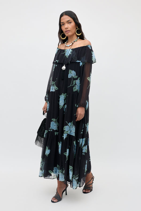 A woman wears the Lela - Peonies Scatter Charcoal, a long, off-the-shoulder black floral dress adorned with blue-green flowers. She pairs it with black strappy heels, large hoop earrings, and carries a black purse while standing against a plain white background.