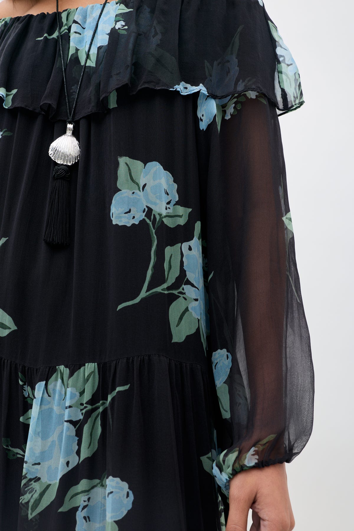 A person wearing the "Lela - Peonies Scatter Charcoal" dress, a sheer black floral piece with blue peonies and green leaves and featuring a ruffled neckline. They accessorize with a silver seashell pendant necklace adorned with a black tassel. Only part of their arm and torso are visible.