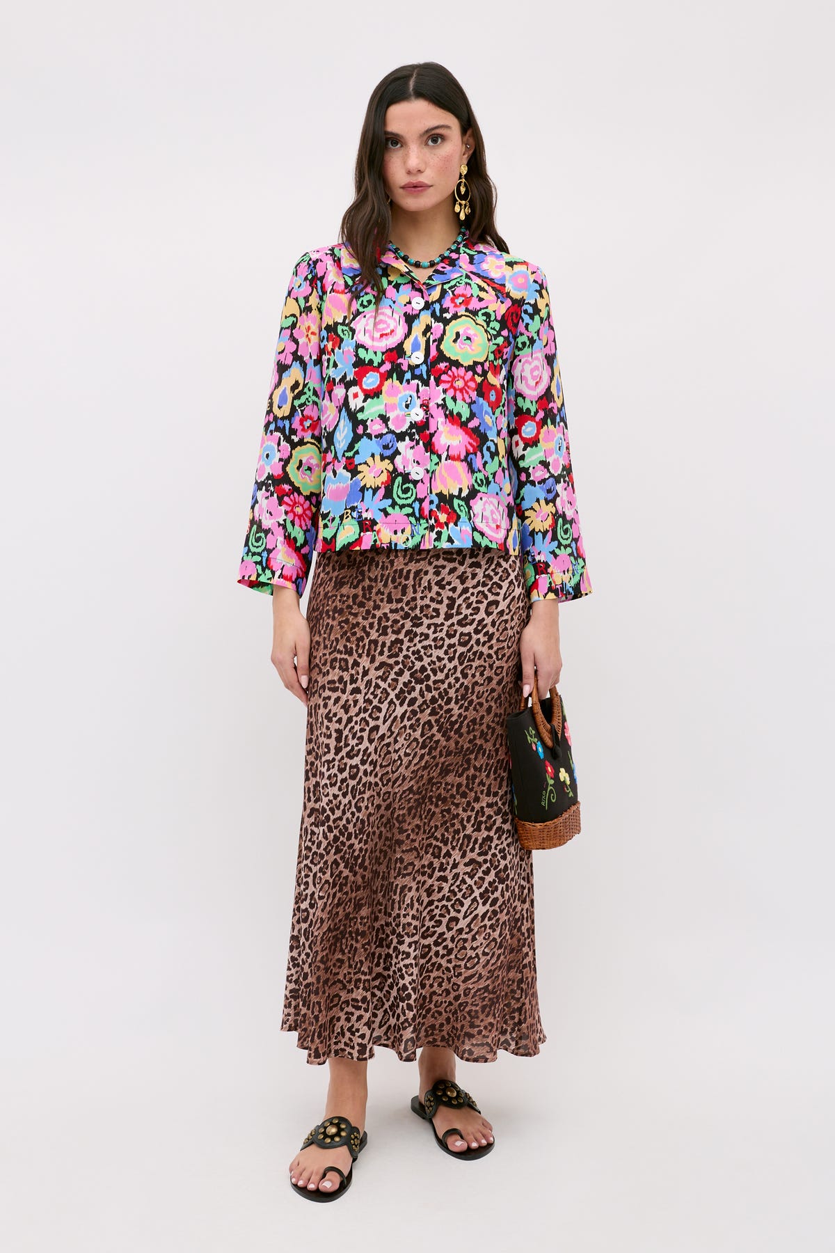 A woman stands wearing a colorful floral blouse, the Kelly - Leopard Brown skirt crafted from luxurious silk crepe de chine featuring a long leopard print design, black sandals, and holding a small handbag. She has long dark hair and wears gold earrings against a plain light background.