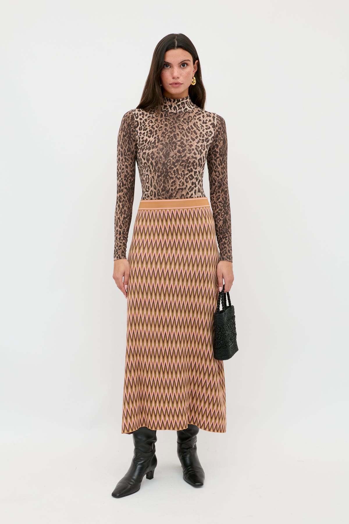 A woman stands against a white background wearing the Brandy - Maeve Jacquard Multi skirt with a leopard print turtleneck, black boots, and holding a small black textured handbag.
