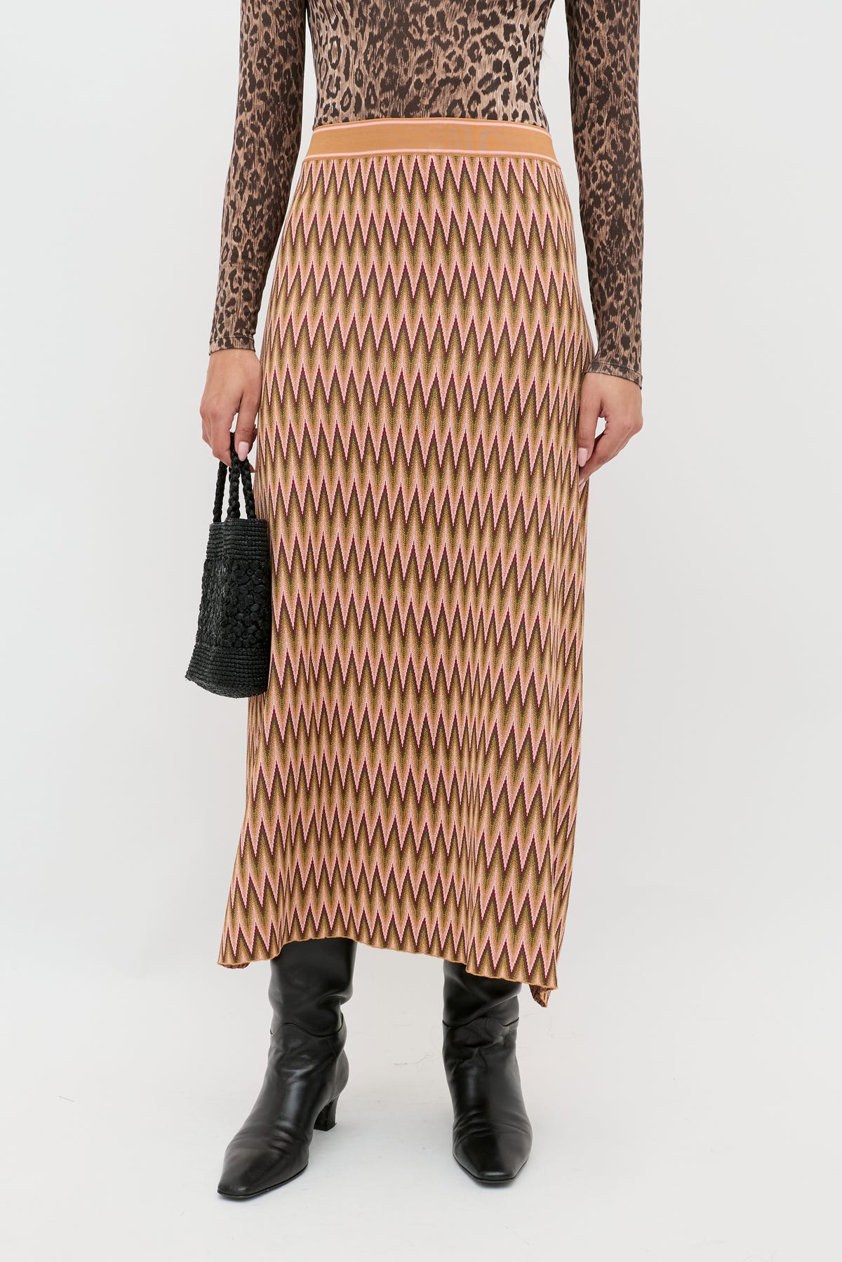 A person wearing the Brandy Maeve Jacquard Multi high-waisted zigzag-patterned midi skirt in tan, pink, and brown with a brown leopard-print long-sleeve top, black knee-high boots, and a small black woven handbag.