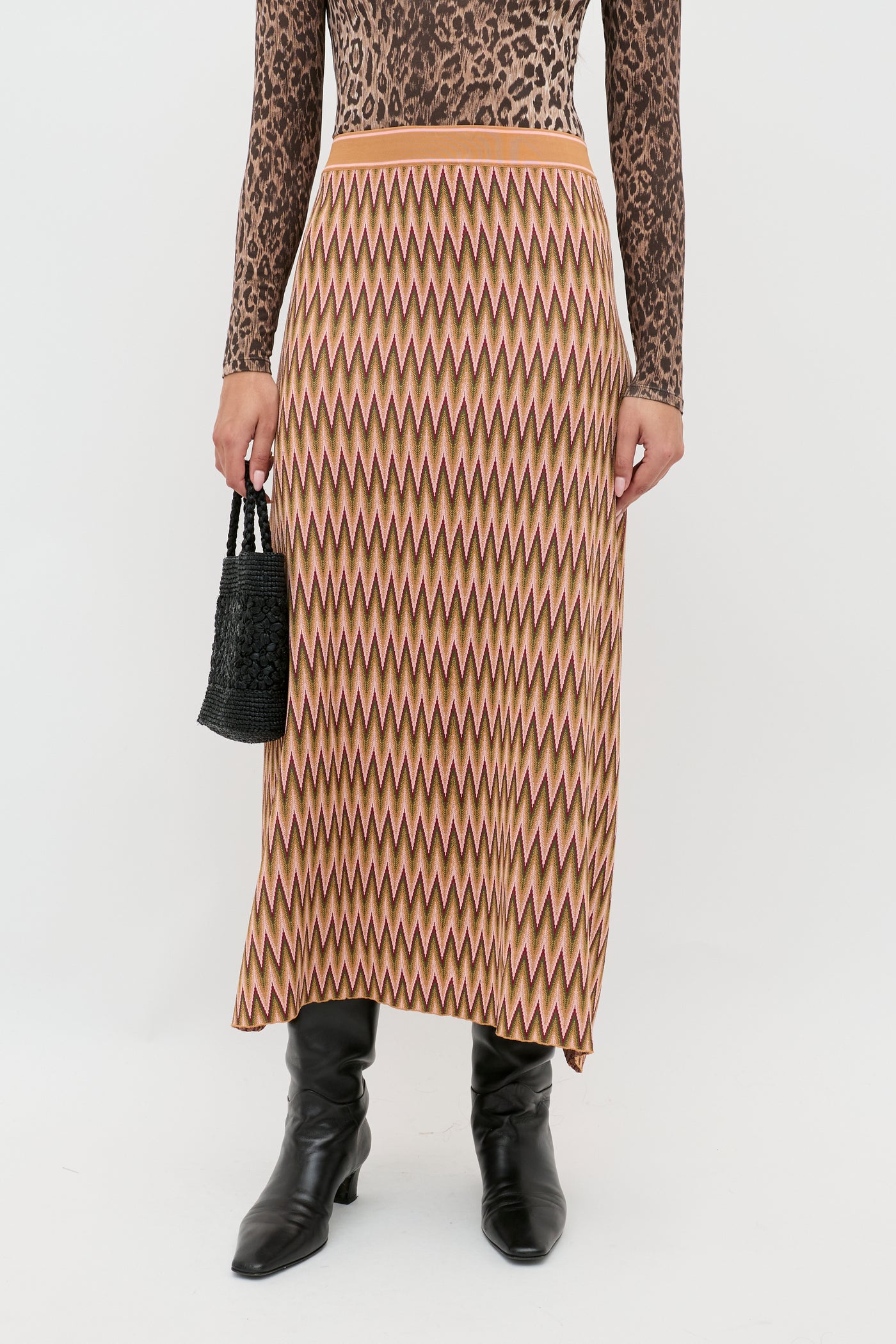 A person wearing the Brandy Maeve Jacquard Multi high-waisted zigzag-patterned midi skirt in tan, pink, and brown with a brown leopard-print long-sleeve top, black knee-high boots, and a small black woven handbag.